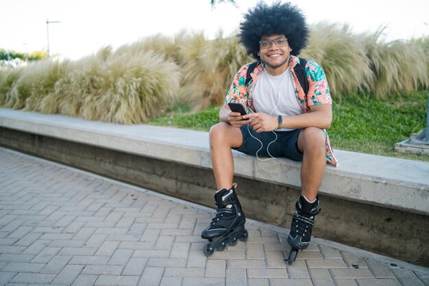 Retrato de joven latino usando su teléfono móvil y usando patines mientras está sentado al aire libre. Concepto deportivo y urbano