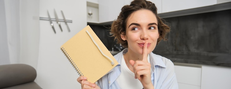 Foto gratuito retrato de una joven linda que sostiene un planificador escribe sus secretos en un diario muestra un gesto de silencio