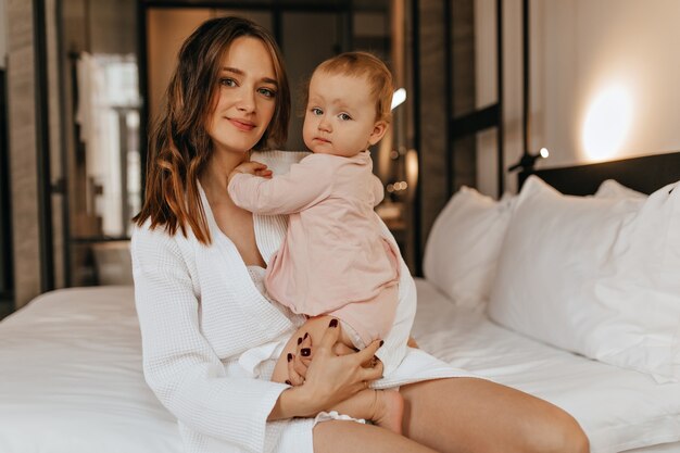 Retrato de joven madre vestida de blanco y su pequeña hija. La mujer y el niño miran a la cámara mientras están sentados en la cama.