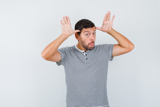 Retrato de joven manteniendo las manos cerca de la cabeza como orejas, sacando la lengua en la camiseta y mirando divertida vista frontal