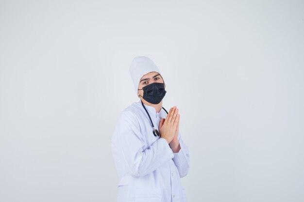 Retrato de joven manteniendo las manos en gesto de oración en uniforme blanco, máscara y mirando esperanzado 