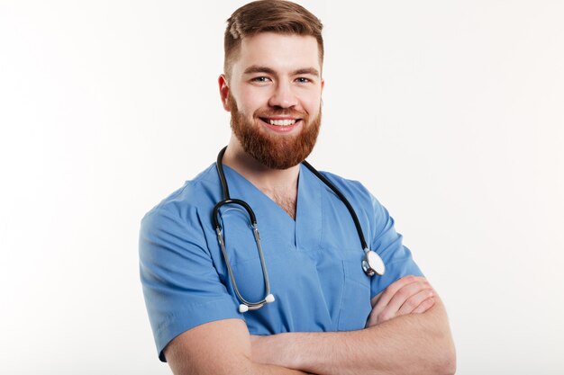 Retrato de un joven médico sonriente con estetoscopio de pie con los brazos cruzados