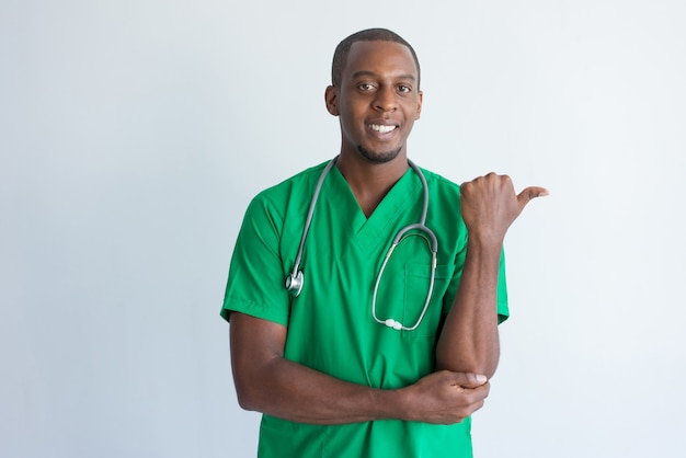 Retrato de joven médico sonriente señalando algo con el pulgar.