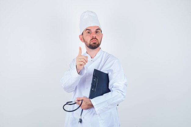 Retrato de joven médico sosteniendo portapapeles, estetoscopio, apuntando hacia arriba en uniforme blanco y mirando inteligente vista frontal