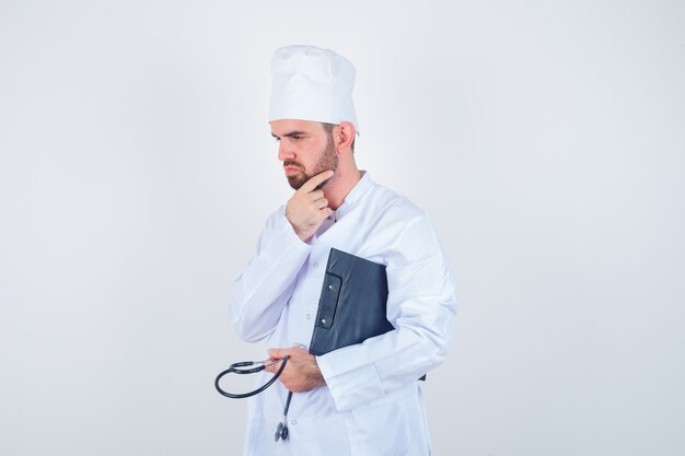 Retrato de joven médico sosteniendo portapapeles, estetoscopio, tocando su barba en uniforme blanco y mirando pensativo vista frontal