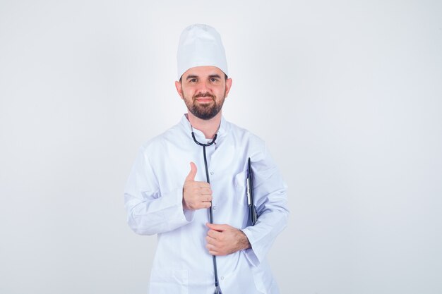 Retrato de joven médico sosteniendo el portapapeles, mostrando el pulgar hacia arriba en uniforme blanco y mirando alegre vista frontal