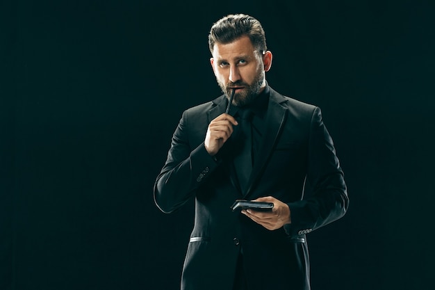 Retrato de un joven de moda con elegante corte de pelo con traje de moda posando sobre pared negra