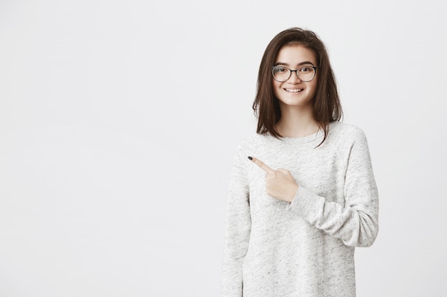 Retrato de joven modelo europeo en gafas apuntando a la izquierda y sonriendo ampliamente