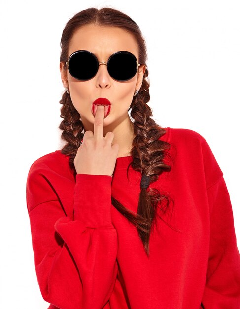 Retrato de joven modelo de mujer sonriente feliz con maquillaje brillante y labios coloridos con dos coletas y gafas de sol en ropa de verano rojo aislado. Lamer el dedo medio, joder signo