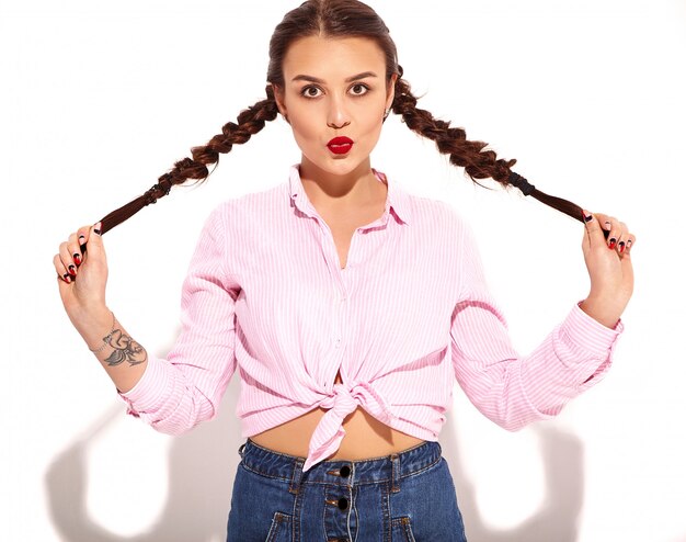 Retrato de joven modelo de mujer sonriente feliz con maquillaje brillante y labios rojos con dos coletas en manos en verano colorida camisa atada de color rosa aislada