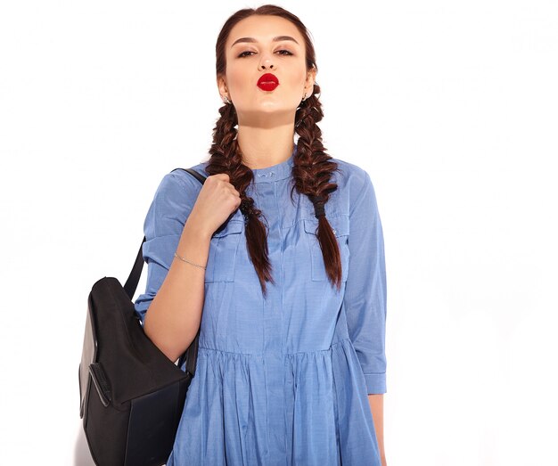 Retrato de joven modelo de mujer sonriente feliz con maquillaje brillante y labios rojos con dos coletas en manos en verano colorido vestido azul y mochila aislado. Dando beso de aire