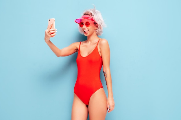 Retrato de joven modelo rubia sonriente en traje de baño de verano traje de baño rojo y gorra de visera transparente Mujer sexy despreocupada divirtiéndose y volviéndose loca Mujer posando cerca de la pared azul en el estudio Tomando selfie
