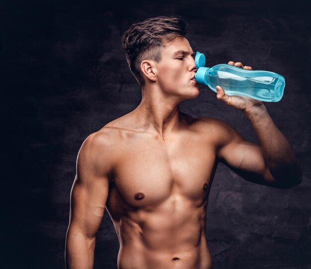 Retrato de un joven modelo sexy sin camisa con un cuerpo musculoso y un corte de pelo elegante, bebe agua refrescante en un estudio. Aislado en un fondo oscuro.