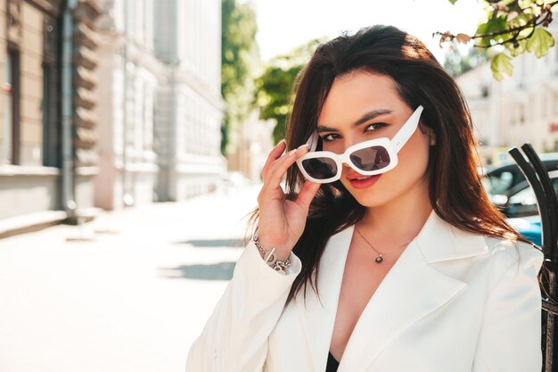 Retrato de joven modelo sexy empresaria moderna. Caliente hermosa mujer en traje blanco posando en el fondo de la calle. Mujer de moda caminando al aire libre con gafas de sol