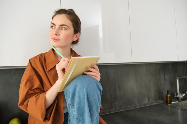 Foto gratuita retrato de una joven moderna con un cuaderno y un bolígrafo sentada en la cocina mirando lejos con