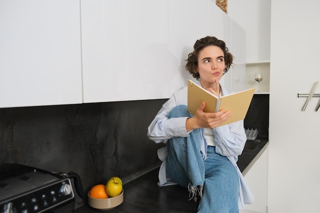 Retrato de una joven moderna leyendo en la cocina sentada en el mostrador y sonriendo estudiando