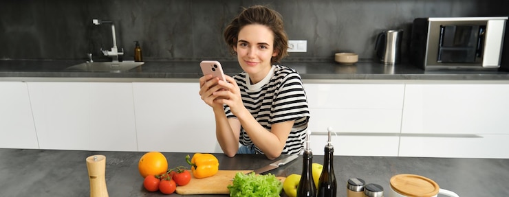 Foto gratuito retrato de una joven moderna pensando qué cocinar sentada en la cocina con un teléfono inteligente y