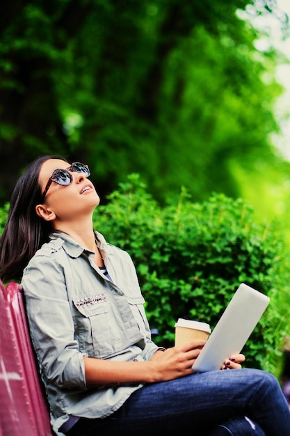 Retrato de una joven morena atractiva con gafas de sol sostiene un tablet Pc y bebe café en un parque verde de verano.