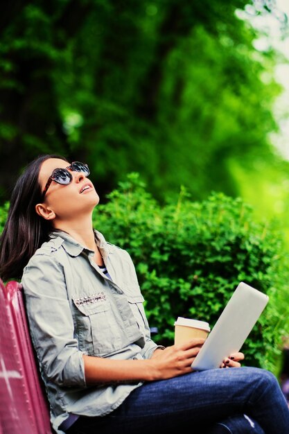 Retrato de una joven morena atractiva con gafas de sol sostiene un tablet Pc y bebe café en un parque verde de verano.