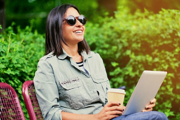 Retrato de una joven morena atractiva con gafas de sol sostiene un tablet Pc y bebe café en un parque verde de verano.