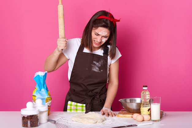 Retrato de joven morena destacó mujer trabajando en la cocina todo el día, preparando pasteles caseros, parece cansado. late en la masa con un rodillo de madera con ira aislado en rosa.