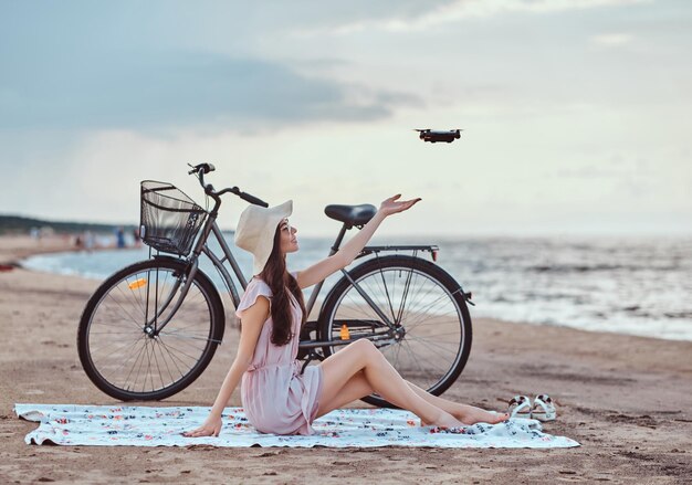 Retrato de una joven morena con gafas de sol y sombrero vestido que disfruta de las vacaciones en la playa, jugando con el cuadricóptero.