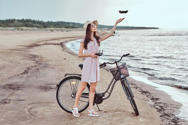 Retrato de una joven morena con gafas de sol y sombrero vestido que disfruta de las vacaciones en la playa, jugando con el cuadricóptero.