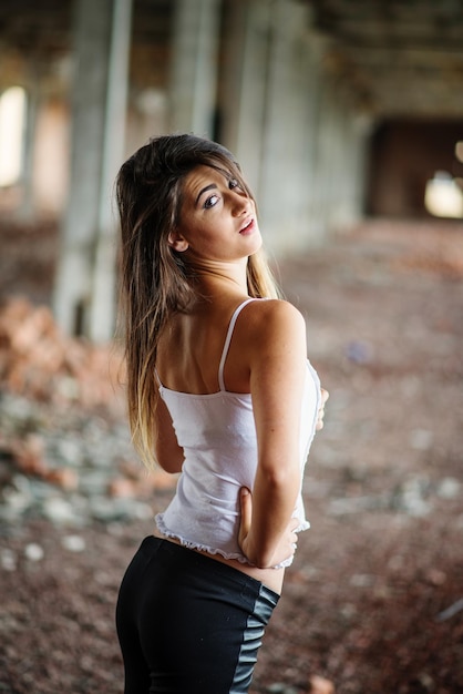 Retrato de una joven morena linda con pantalones de cuero negro y blusa blanca posada en un lugar abandonado