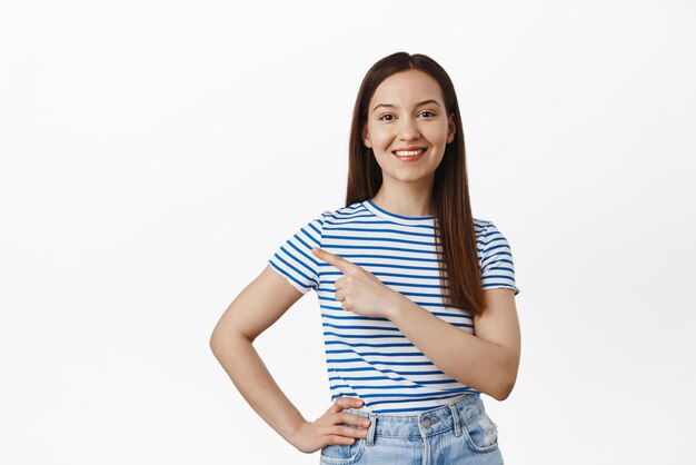 Retrato de una joven morena que señala con el dedo a la izquierda mostrando el punto de promoción de venta en el logotipo sonriendo feliz recomendar hacer clic en el enlace de pie sobre fondo blanco