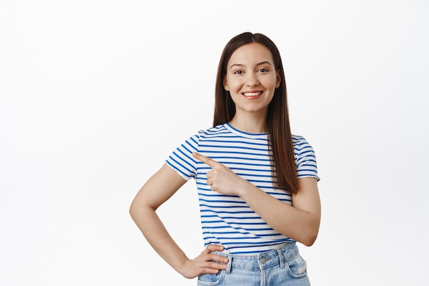 Retrato de una joven morena que señala con el dedo a la izquierda mostrando el punto de promoción de venta en el logotipo sonriendo feliz recomendar hacer clic en el enlace de pie sobre fondo blanco