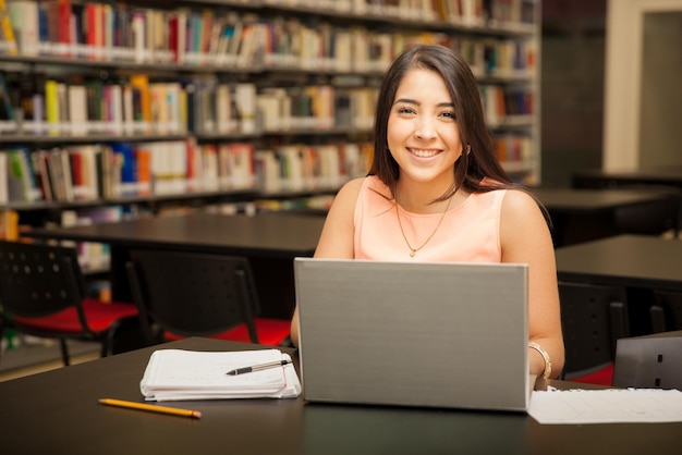 Retrato de una joven morena usando una computadora portátil para el trabajo escolar en la biblioteca y sonriendo