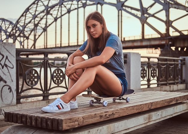 Retrato de una joven morena vestida con una camiseta y pantalones cortos sentada en una patineta al fondo del viejo puente al atardecer.