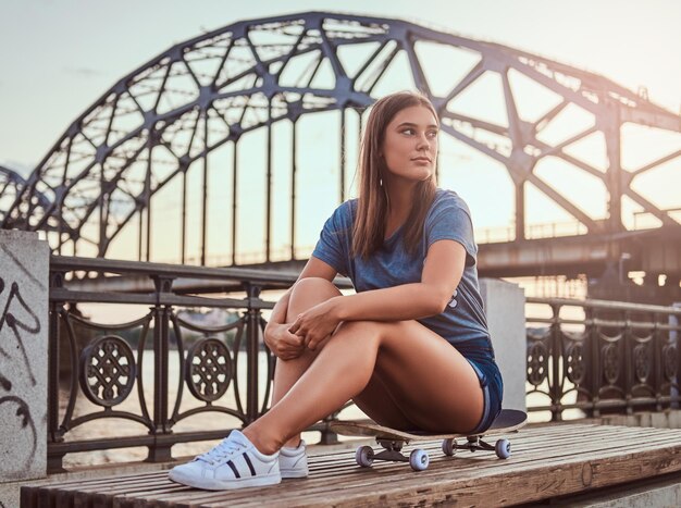 Retrato de una joven morena vestida con una camiseta y pantalones cortos sentada en una patineta al fondo del viejo puente al atardecer.