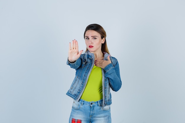 Retrato de joven mostrando gesto de rechazo, manteniendo la mano en el pecho con traje de mezclilla y mirando reacia vista frontal