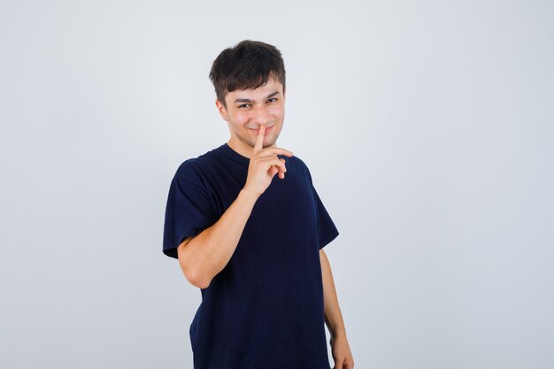 Retrato de joven mostrando gesto de silencio en camiseta negra y mirando confiado vista frontal