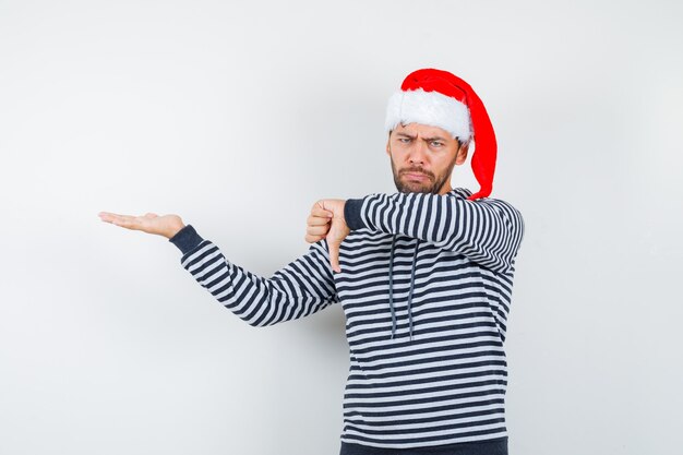 Retrato de joven mostrando el pulgar hacia abajo, extendiendo la palma a un lado en la sudadera con capucha, gorro de Papá Noel y mirando disgustado