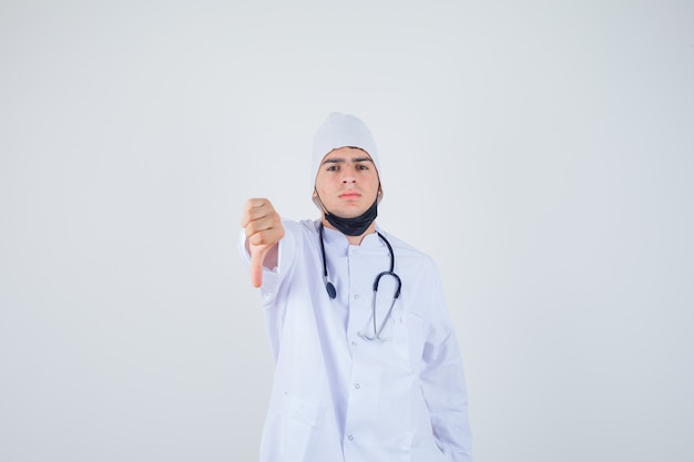 Retrato de joven mostrando el pulgar hacia abajo en uniforme blanco, máscara y mirando disgustado