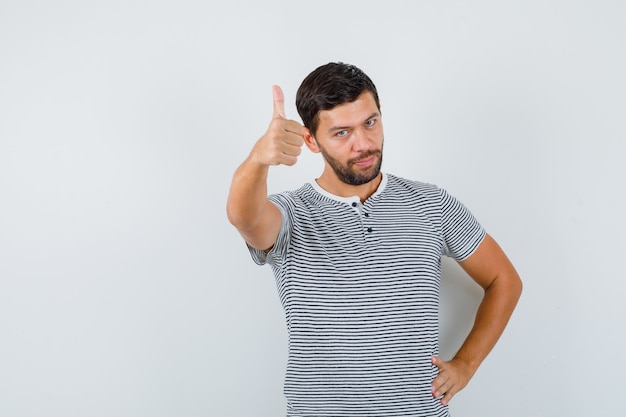 Retrato de joven mostrando el pulgar hacia arriba en camiseta y mirando complacido vista frontal