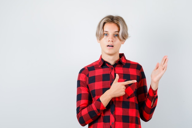 Foto gratuita retrato de joven muchacho adolescente apuntando a su mano levantada en camisa a cuadros y mirando sorprendido vista frontal