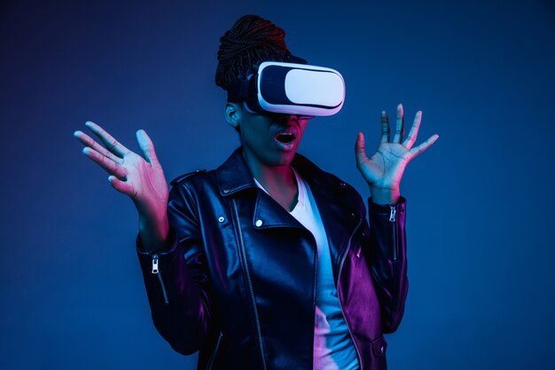 Retrato de joven mujer afroamericana jugando en gafas de realidad virtual en luz de neón en azul.