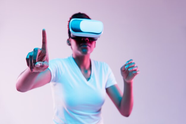 Retrato de joven mujer afroamericana jugando en gafas VR en luz de neón en gradiente.