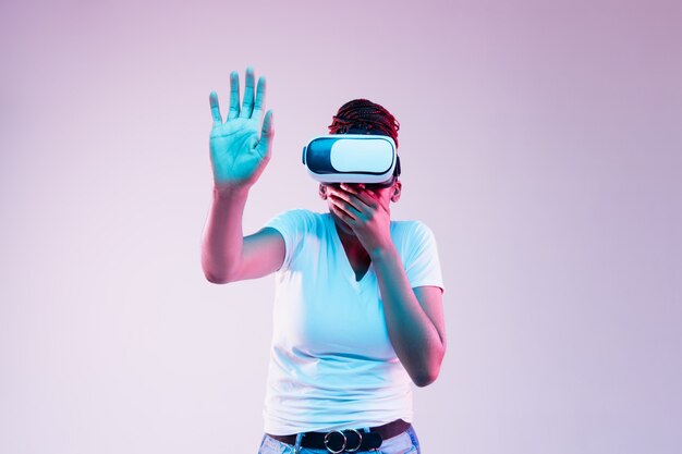 Retrato de joven mujer afroamericana jugando en gafas VR en luz de neón sobre fondo degradado. Concepto de emociones humanas, expresión facial, tecnología y aparatos modernos. Toca algo.