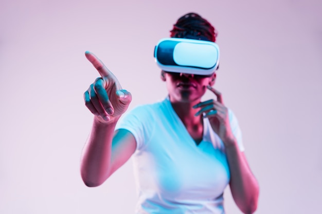 Retrato de joven mujer afroamericana jugando en gafas VR en luz de neón sobre fondo degradado