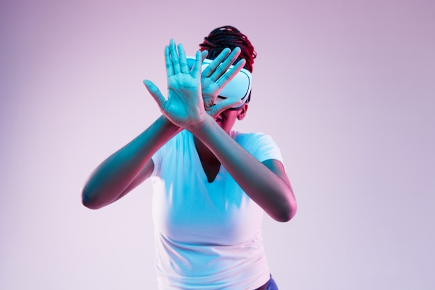 Retrato de joven mujer afroamericana jugando en gafas VR en luz de neón sobre fondo degradado