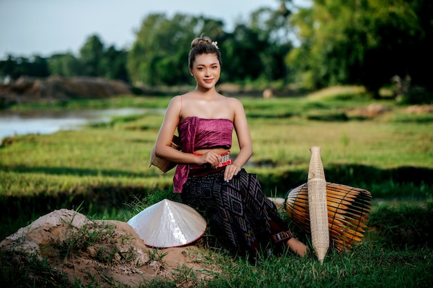 Retrato joven mujer bonita asiática en hermosa ropa tradicional tailandesa en el campo de arroz, ella sentada cerca de equipos de pesca