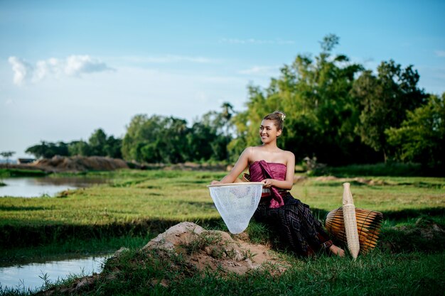 Retrato joven mujer bonita asiática en hermosa ropa tradicional tailandesa en el campo de arroz, ella sentada cerca de equipos de pesca