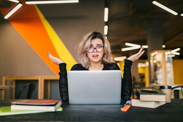 Retrato de joven mujer bonita con expresión de la cara con calcetines, sentado en la mesa trabajando en la computadora portátil en la oficina de co-trabajo, con gafas, estrés en el trabajo, emoción divertida, estudiante en el aula, frustración