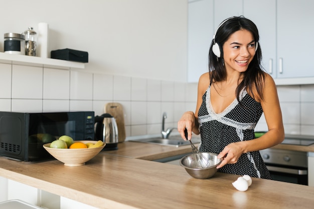 Retrato de joven mujer bonita morena cocinando huevos revueltos en la cocina por la mañana, sonriente, feliz, ama de casa positiva, estilo de vida saludable