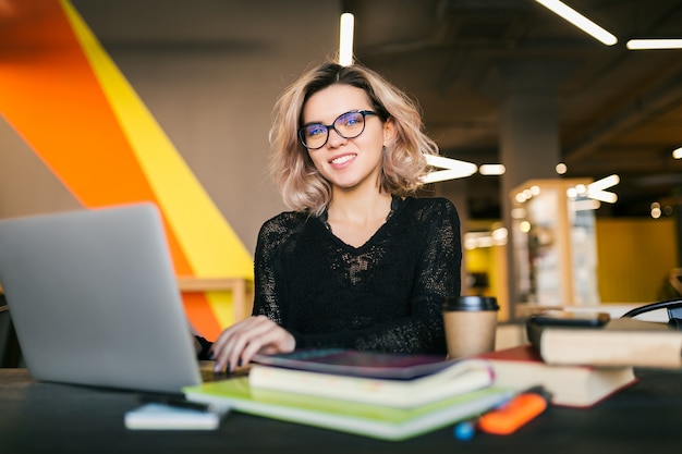 Retrato de joven mujer bonita sentada a la mesa en camisa negra trabajando en la computadora portátil en la oficina de trabajo conjunto, con gafas, sonriendo, feliz, positivo