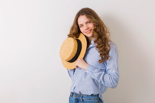 Retrato de joven mujer bonita con sombrero de paja jeans camisa de algodón azul posando en la pared blanca